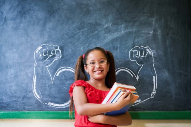 Concept on blackboard at school. Intelligent and successful hispanic girl in class. Portrait of female child smiling, looking at camera, holding books against drawing of muscles on blackboard                     (Concept on blackboard at school. Intel